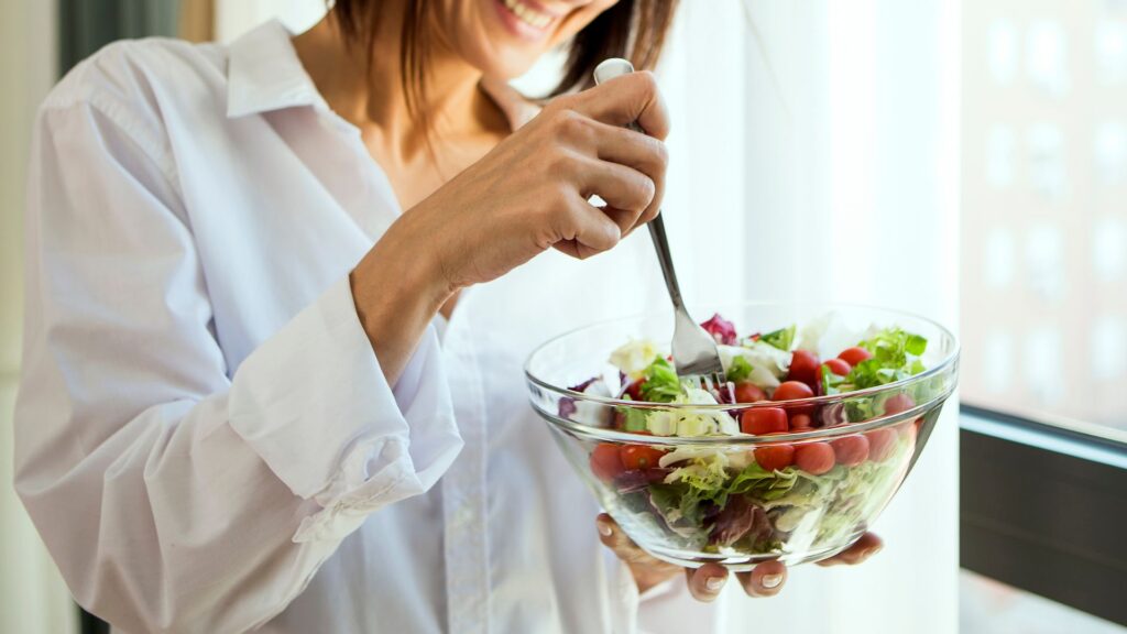Mujer sonriendo mientras sostiene y come una ensalada fresca de vegetales en un ambiente iluminado.